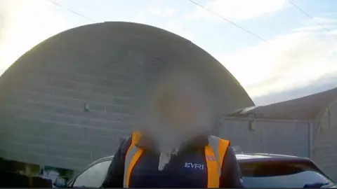 一名面部模糊的女子身穿橙色高可见度背心和蓝色抓绒衣。背景是建筑物屋顶的弧形。