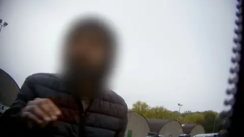 一名面部模糊的男子身穿黑色防风外套。背景是灰色天空和远处的树木。照片为卧底拍摄，右侧可见部分拉链。