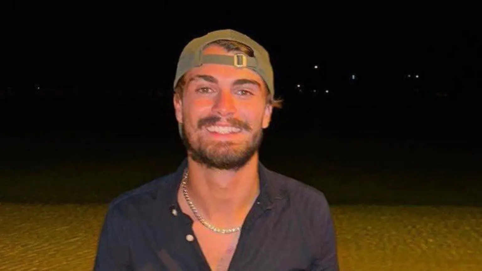 Rockdale Ilinden FC Dan Elkayam smiling against a black background. He is a young man with a short brown beard and moustache, and wears a green sunhat backwards on his head. He is wearing a blue buttoned shirt and a shell necklace.